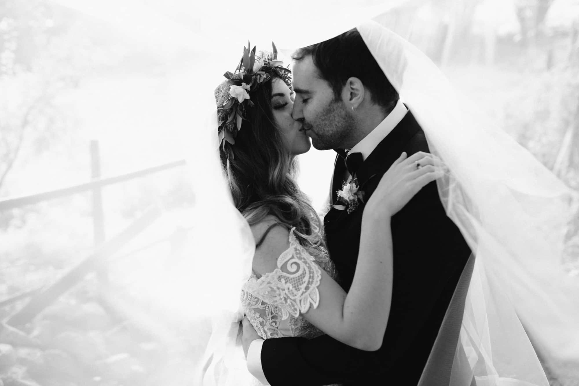 A bride and groom share a kiss under a flowing veil. The bride wears a floral crown and lace dress, whilst the groom is in a suit. The image is in black and white, creating a soft, romantic atmosphere.