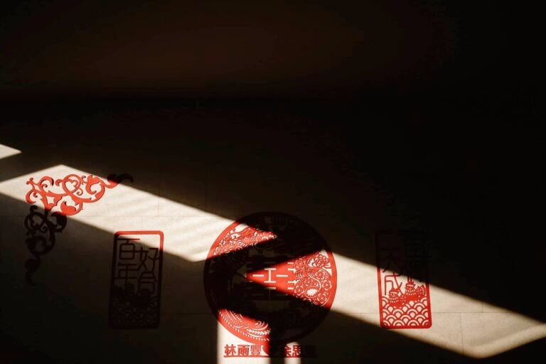 A beam of sunlight cuts across a wall decorated with intricate red Chinese paper cuttings, casting shadows and highlighting traditional designs and Chinese characters—evoking the joyful spirit of a Hong Kong wedding.