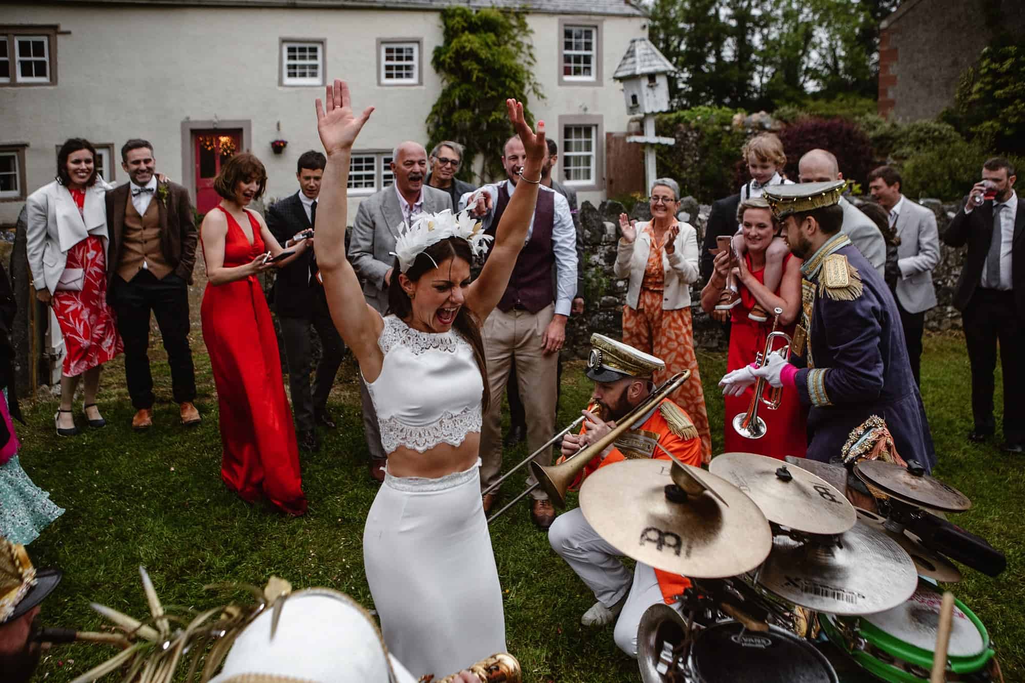A joyful bride in a white two-piece dress raises her arms, celebrating with guests and a lively band performing outdoors in front of a stone house. Guests smile, laugh, and watch the festive scene.