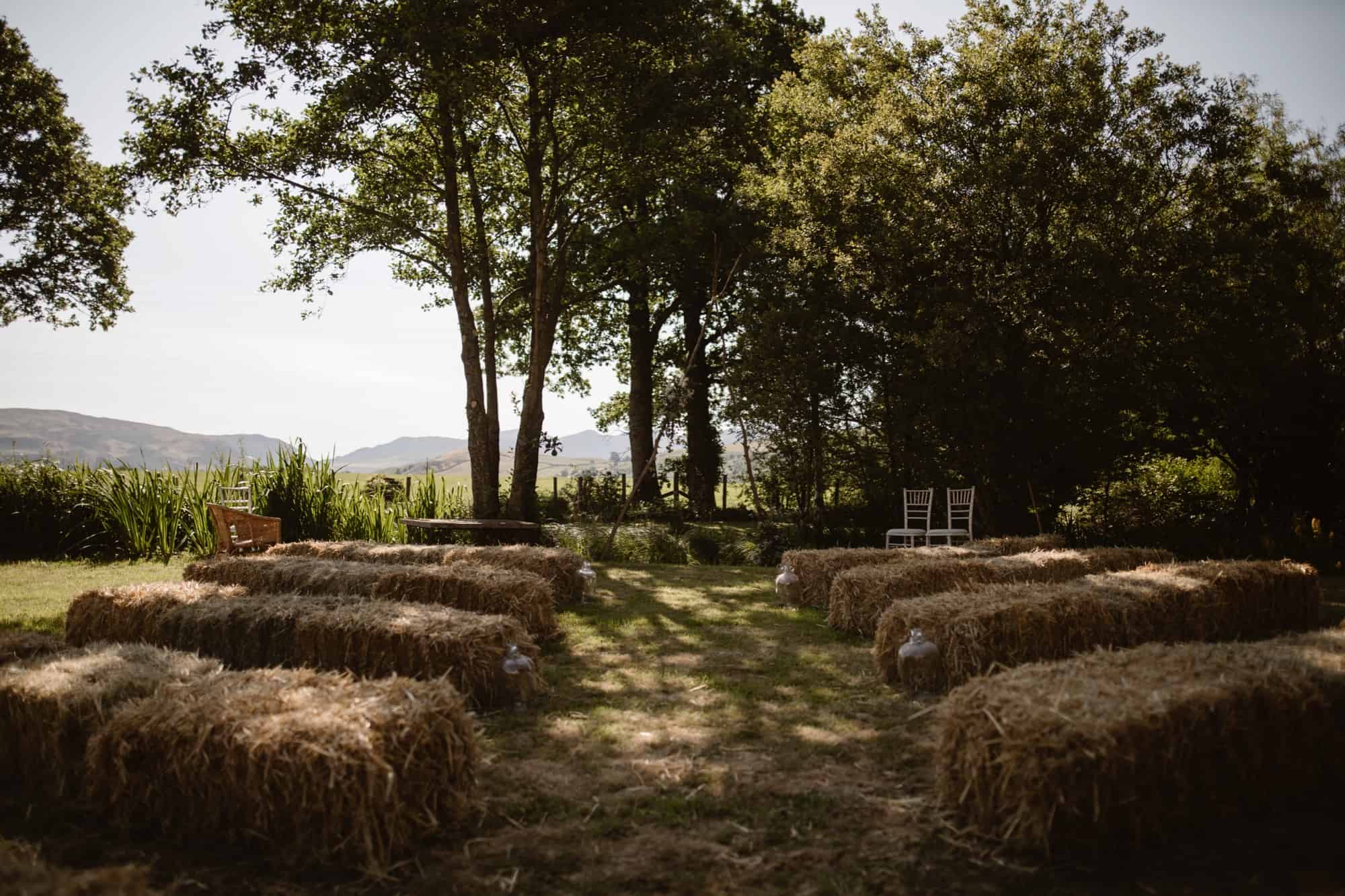 Rows of hay bales arranged as seating face a small open area under trees at Low Hall, creating a rustic outdoor setting with two chairs at the front and hills visible in the background.