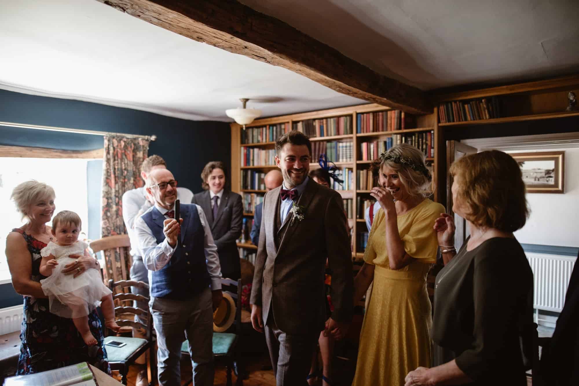 A group of people, including a couple dressed for a special occasion, smile and laugh together inside the cosy Low Hall with bookshelves and wooden beams. One woman holds a baby whilst others stand nearby, enjoying the moment.