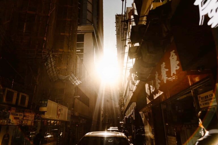 Sunlight shines brightly between tall buildings on a narrow city street, casting long shadows. Cars are parked along the sides, and signs with Chinese characters hint at a bustling, urban Hong Kong Wedding scene unfolding amidst the vibrant city life.