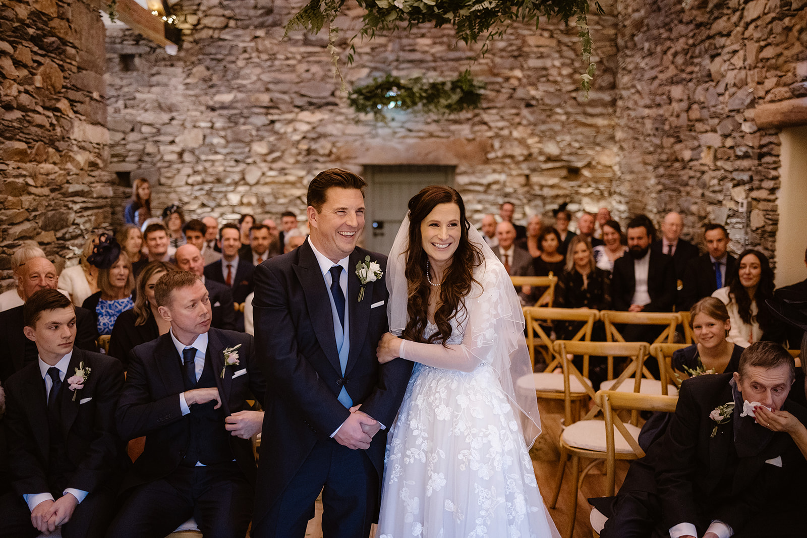 A bride and groom stand smiling at the front of a rustic stone-walled venue at their Town Head Estate Wedding, surrounded by happy, seated guests. The bride wears a white dress and veil; the groom is in a dark suit with a buttonhole.