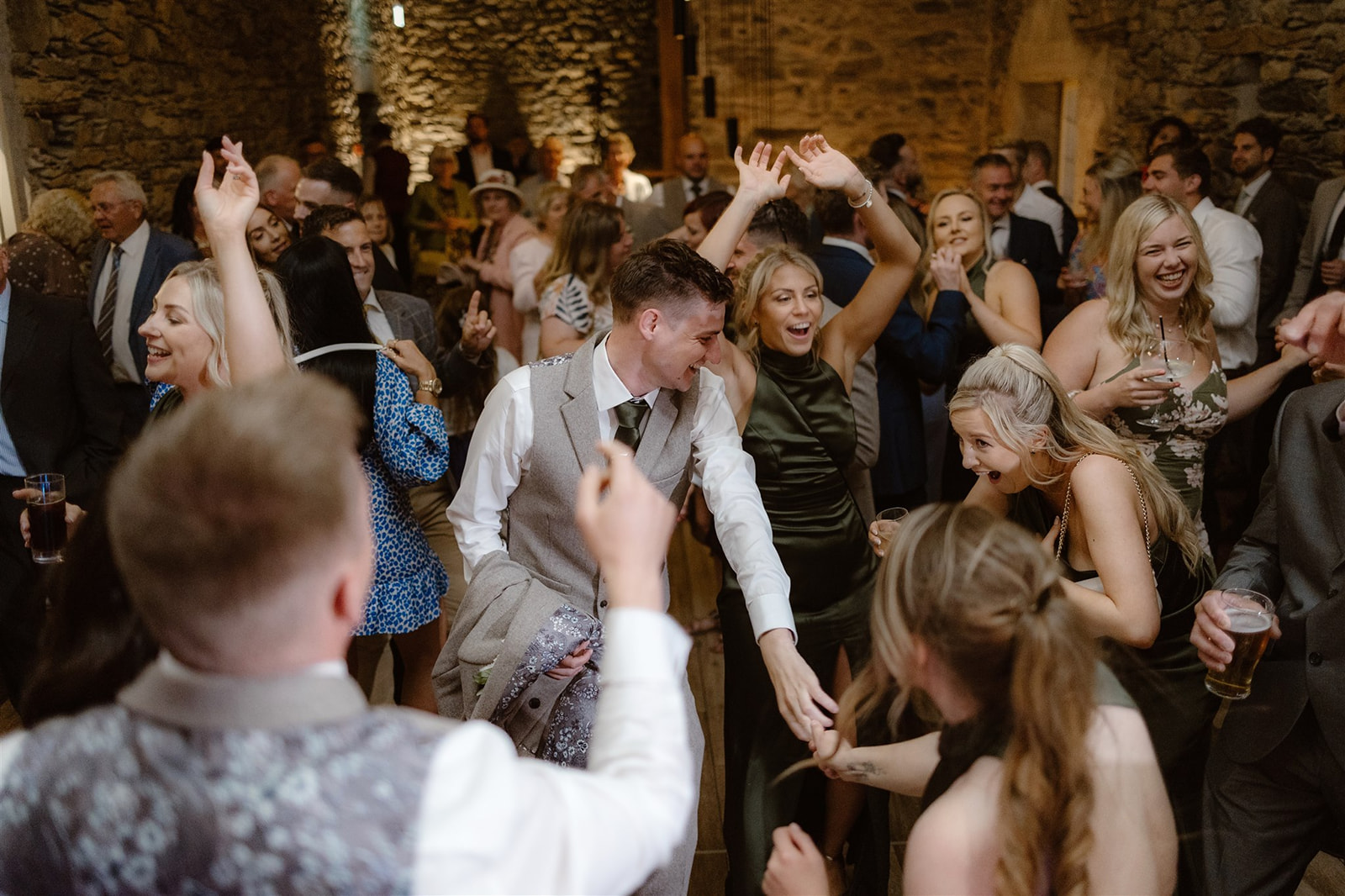 A lively group of people dressed in formal attire dance and celebrate at a Town Head Estate Wedding, smiling and raising their hands in the air against the venue’s charming stone walls.
