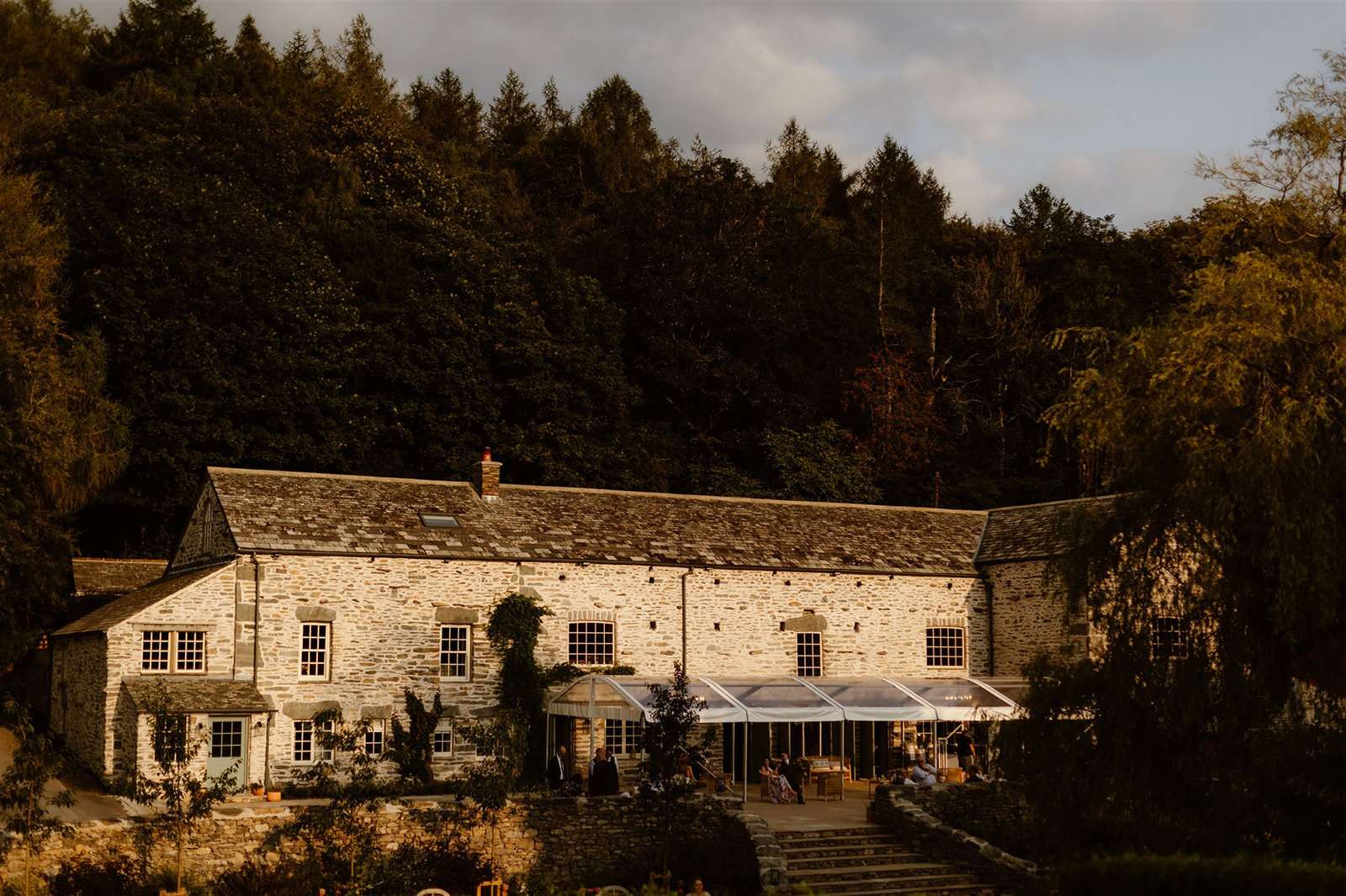 A rustic stone building with large windows stands before a forest of tall trees at Town Head Estate. A glass-covered patio with people seated beneath it is attached to the building, bathed in warm, late afternoon sunlight.