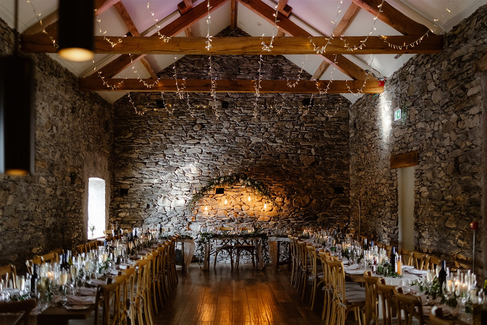 Rustic Town Head Estate banquet hall with stone walls, wooden beams, and fairy lights. Two long dining tables set with glassware, candles, and greenery lead to a top table beneath an arched greenery arrangement—a perfect Town Head Estate Wedding setting.