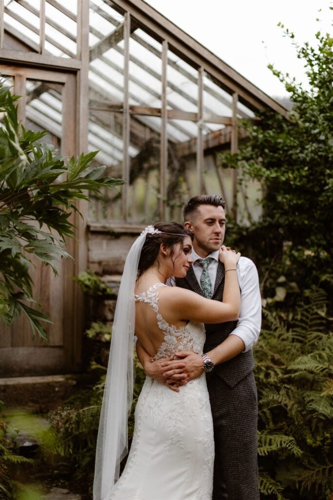 Bride and groom embrace by a greenhouse.