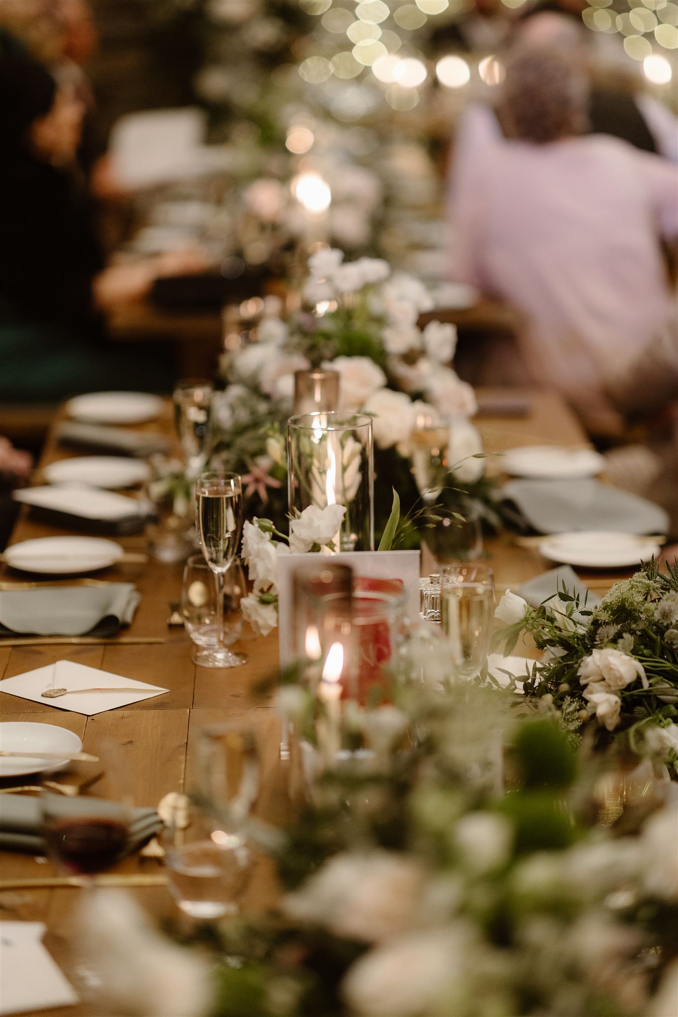 A long wooden table set for a formal event at Town Head Estate, decorated with flowers, candles, plates, and glasses. The softly blurred background features people and twinkling lights, creating a warm, festive atmosphere perfect for a Town Head Estate Wedding.