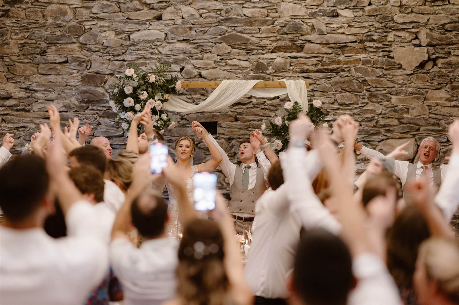 A bride and groom stand before a stone wall at Town Head Estate, smiling and raising their hands joyfully with wedding guests, who also have their arms raised. Some guests hold up phones to capture this unforgettable Town Head Estate Wedding moment.