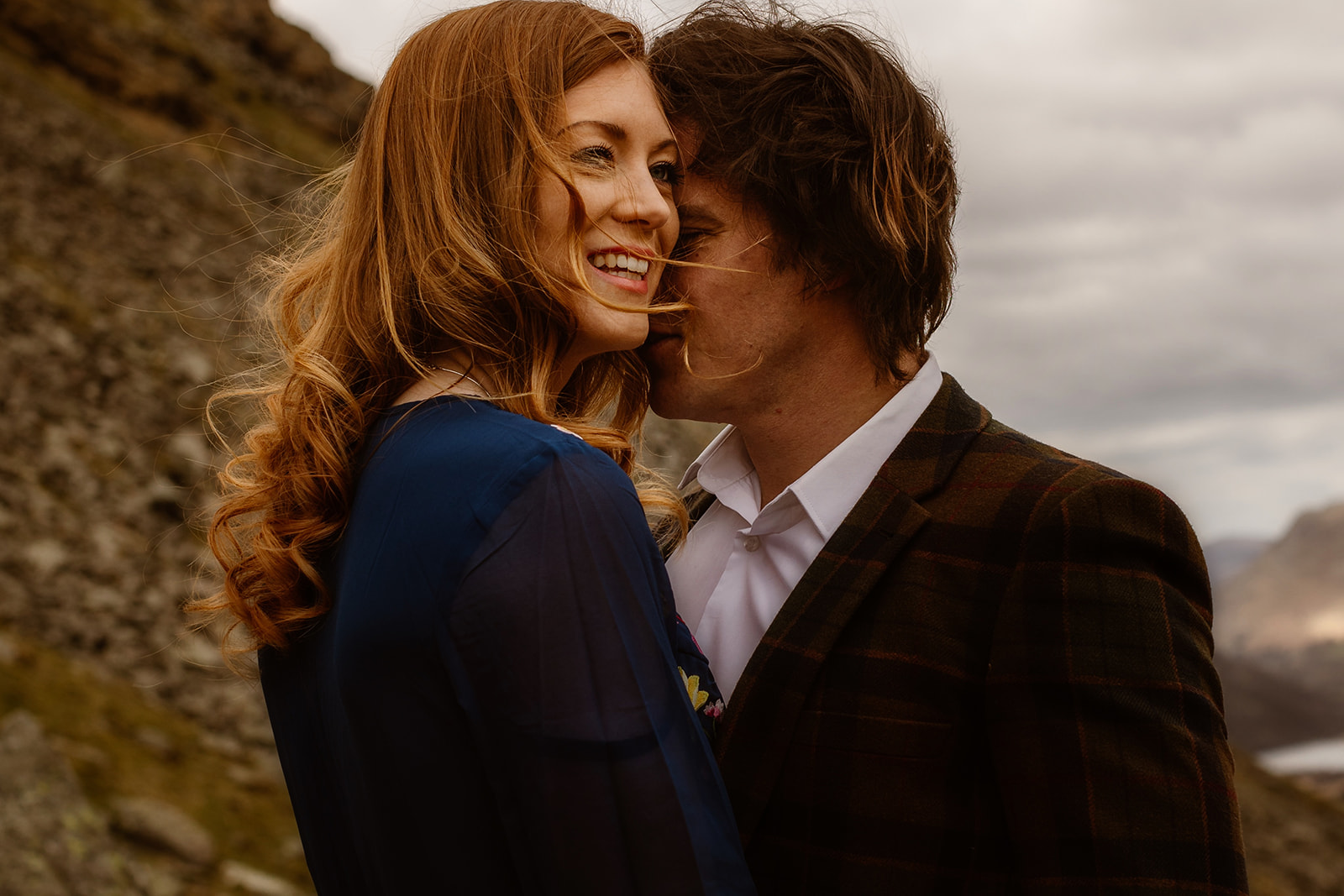 A smiling woman with long, wavy red hair stands next to a man with brown hair in a checked jacket. They are outdoors in a mountainous landscape under a cloudy sky, capturing ideas on how to plan the perfect engagement photoshoot. The man is leaning in close to her.