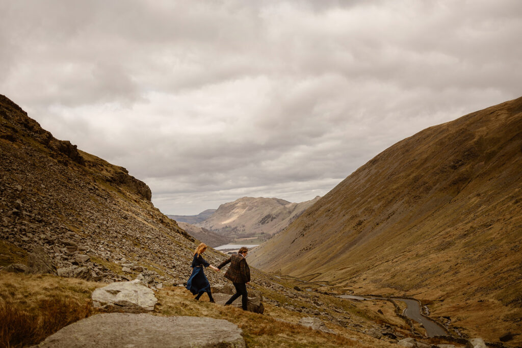 A man leads a woman by her hand down a rocky hillside, with mountainous views and a lake in the background