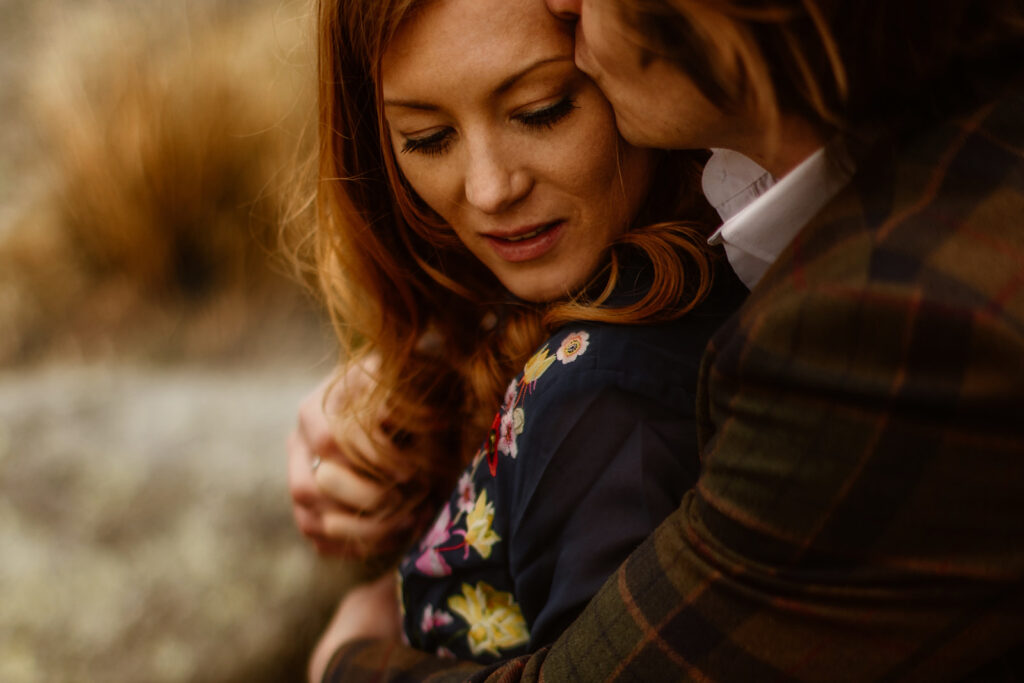 A woman is embraced by her fiancé in a plaid jacket, who is kissing her head in an intimate pre-wedding shoot.