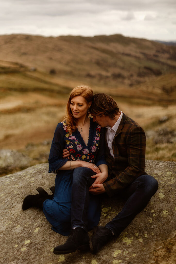 A woman and man sit closely together on a large rock in a hilly landscape, perfect for an engagement shoot. The man rests his head on the woman's shoulder as they hold hands. She wears a blue dress while he wears a brown jacket, creating a picturesque scene for any pre-wedding planning guide.