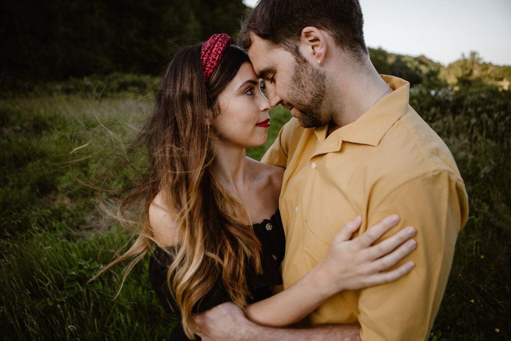 A woman in a black off-shoulder top and a red headband embraces a man in a yellow shirt as they gaze into each other's eyes in a field with green foliage.