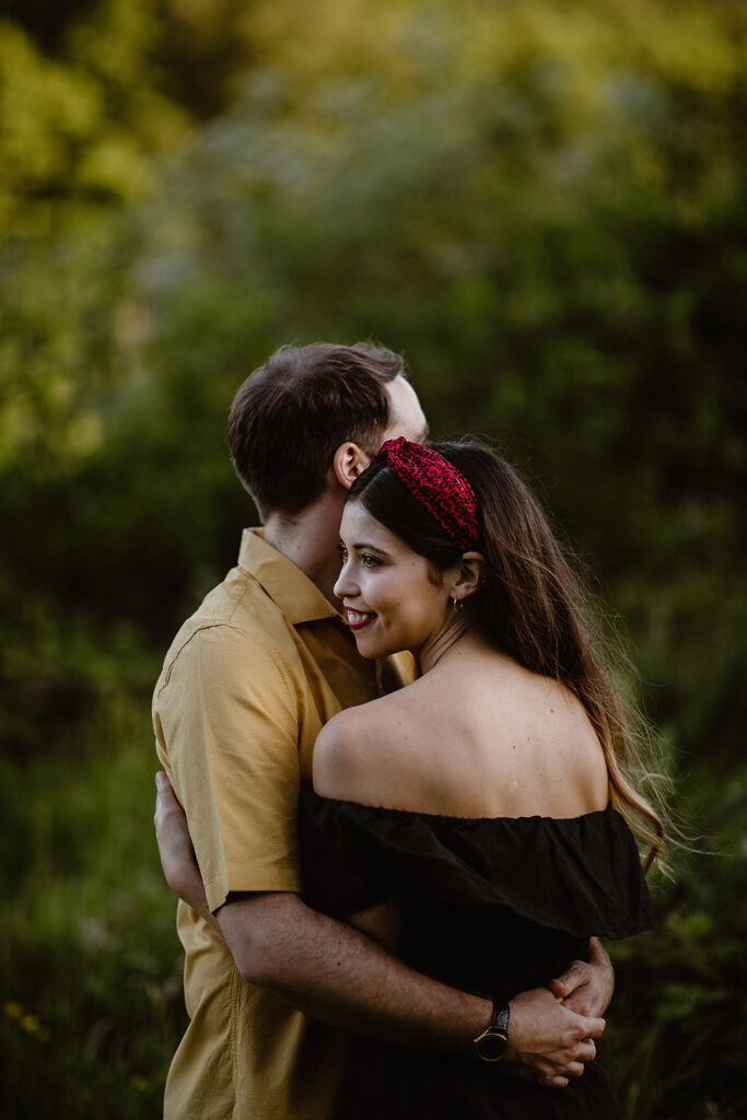 A woman in a black off-shoulder top and a red headband looks into the distance whist she embraces a man in a yellow shirt in a field with green foliage.