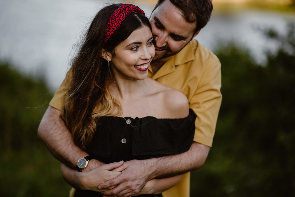 A man in a yellow shirt embraces a woman in a black off-shoulder top, both smiling amidst lush greenery.