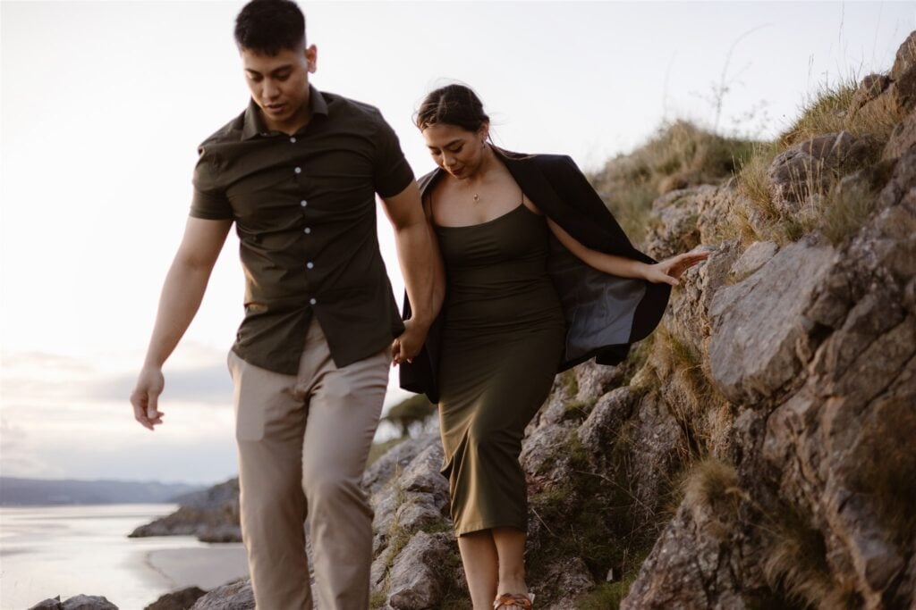 A man and a woman holding hands while carefully walking down rocky terrain near a body of water, with the sky visible in the background