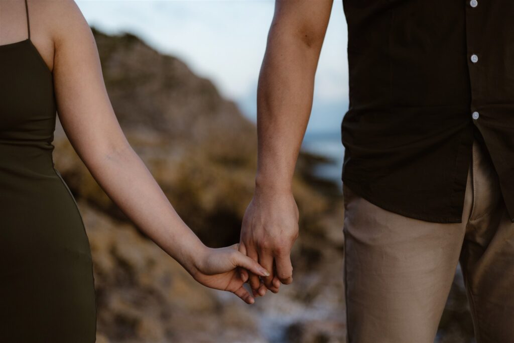A closeup of a couple holding hands with a rocky seaside backdrop behind them - the perfect engagement shoot location