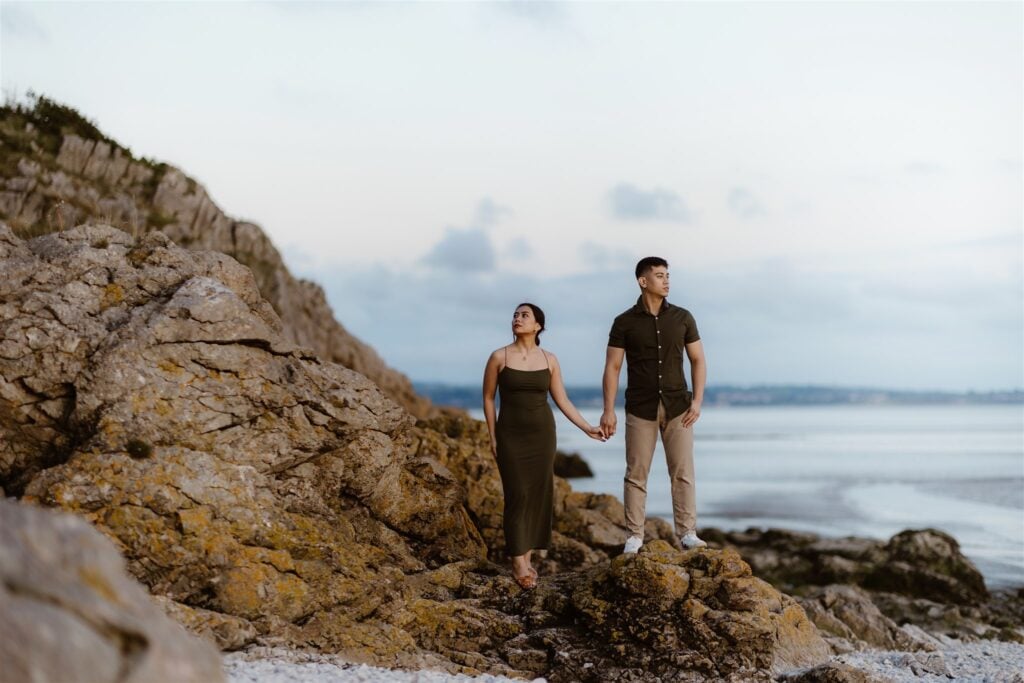 Couple on engagement shoot stand on rocky terrain by the seaside, holding hands and looking into the distance. The sky is overcast and the sea is visible in the background.