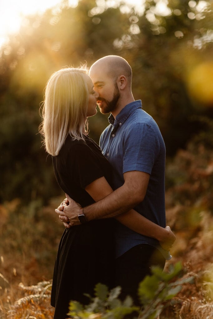 An engaged couple embrace outdoors at sunset during their engagement shoot. The woman is leaning into the man to kiss him.