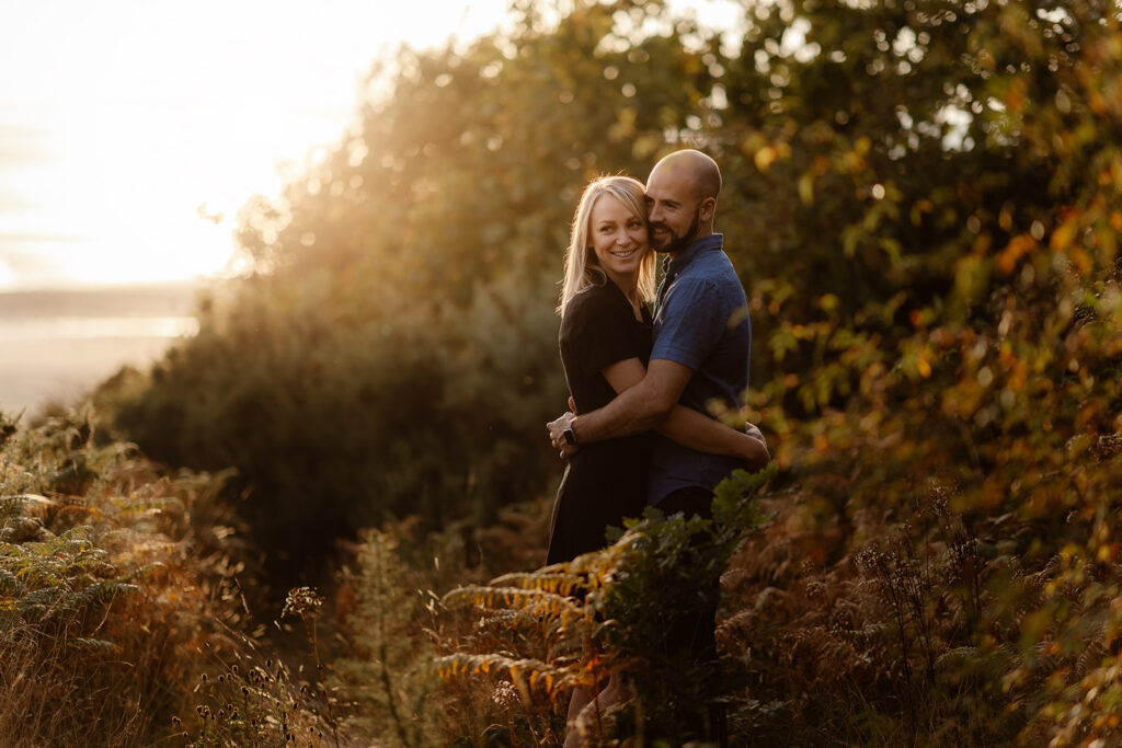 A man and woman embrace looking outdoors at sunset during their engagement shoot. They are lookking off into the distance.