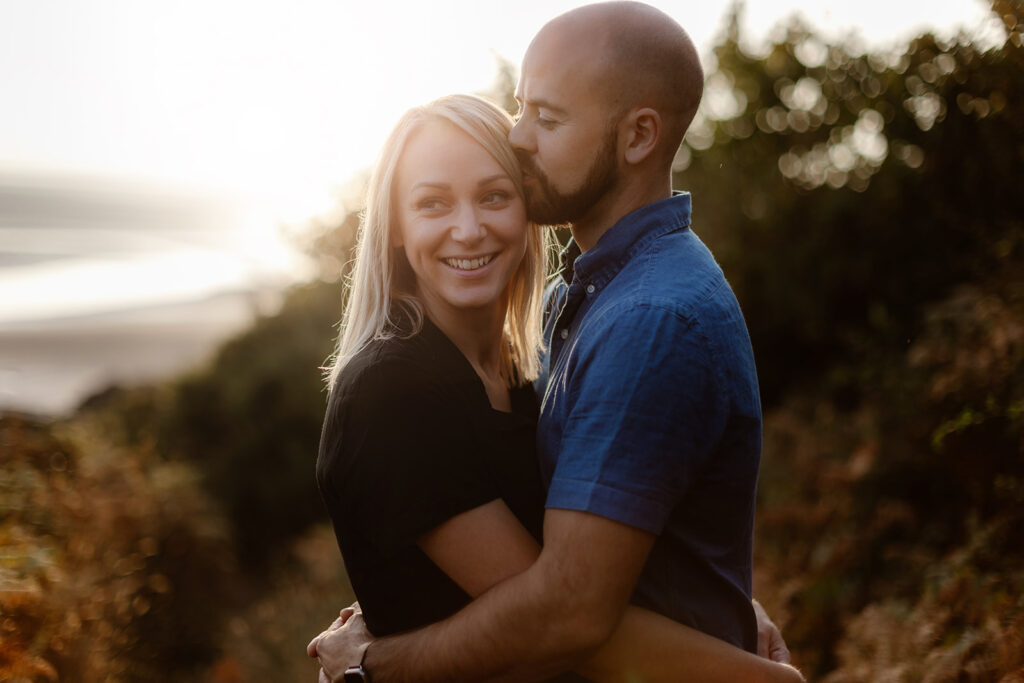 A man and woman embrace outdoors at sunset during their engagement shoot. The man kisses the woman's temple as she smiles, with trees and a blurred background behind them.