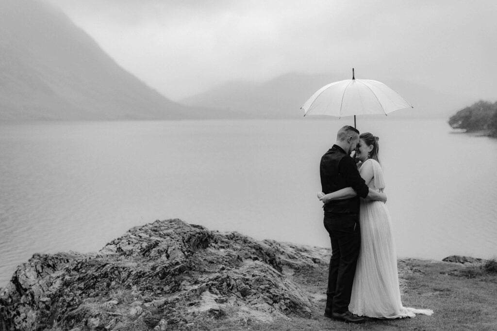Bride & Groom stood next to a lake under an umbrella, kissing during their elopement
