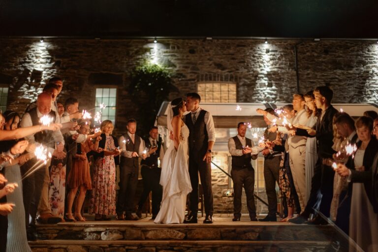 Bride & Groom kissing as guests look on, holding sparklers at night time