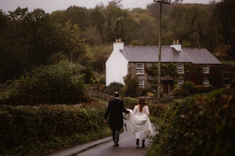 Bride wearing wellingtons and holding her skirt up walking down country road with husband