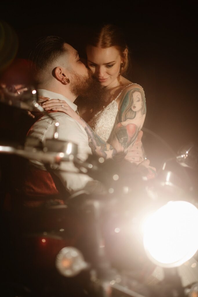 Bride sat on her husband's lap with a motorbike in the foreground during their small intimate wedding