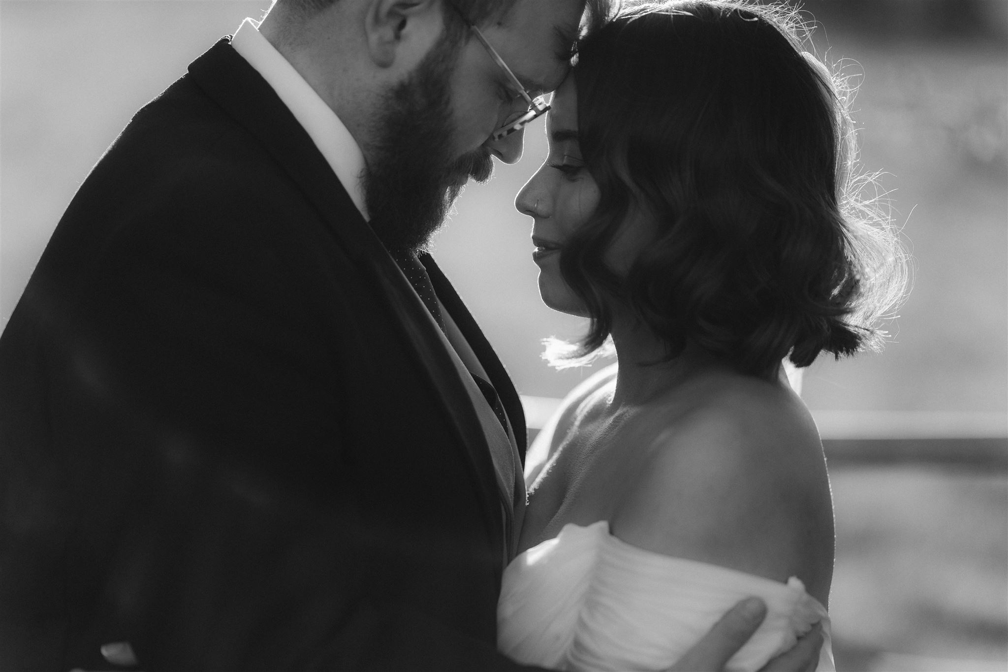 A black and white photo of a couple embracing closely, foreheads touching, with soft natural light highlighting their faces. The woman wears an off-shoulder dress and the man wears a suit and glasses.