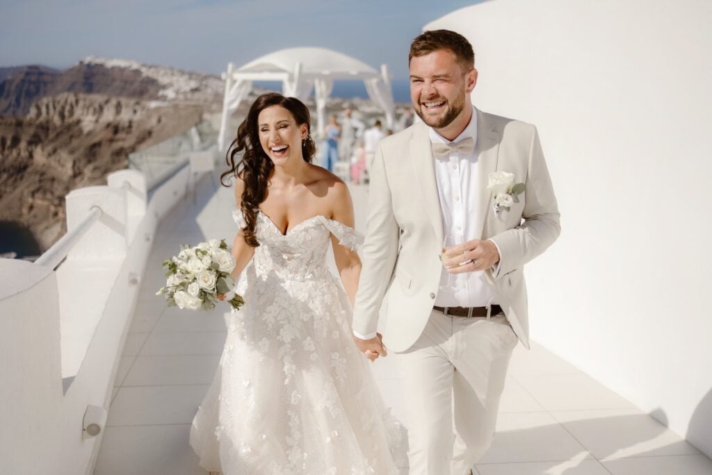 A joyful bride and groom, dressed in elegant wedding attire, hold hands and smile as they walk outdoors on a sunny day, surrounded by white architecture and scenic mountain views—capturing the essence of Destination Wedding Photography.