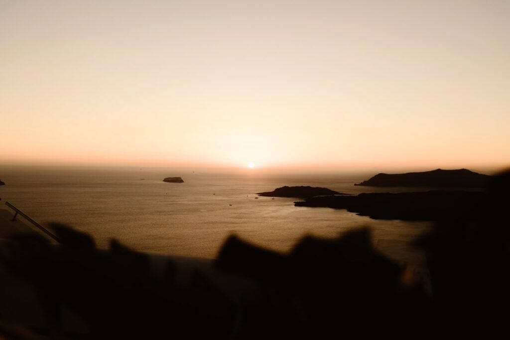 Golden sunset over a calm sea with distant islands and silhouettes of hills on the horizon; the sky is softly lit with warm tones—a perfect scene for destination wedding photography.