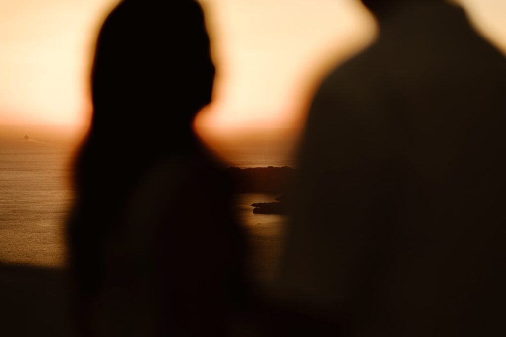 Silhouettes of two people facing each other with a blurred sea and island in the background at sunset, creating a warm, romantic atmosphere—perfect inspiration for destination wedding photography.