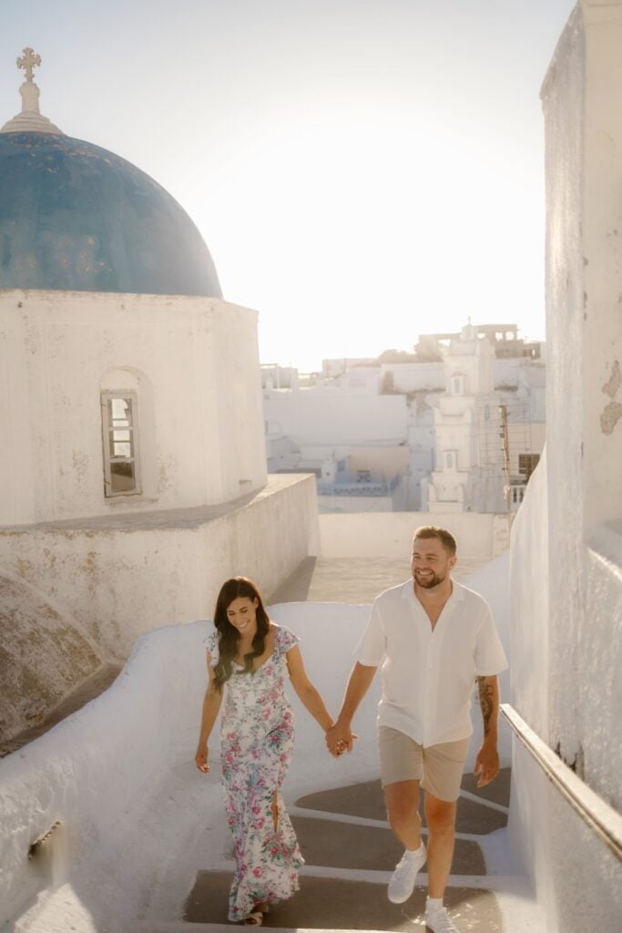 A couple holding hands and smiling as they walk up white stairs in a sunlit, whitewashed town with a blue-domed building—perfect for Destination Wedding Photography in stunning Santorini, Greece.
