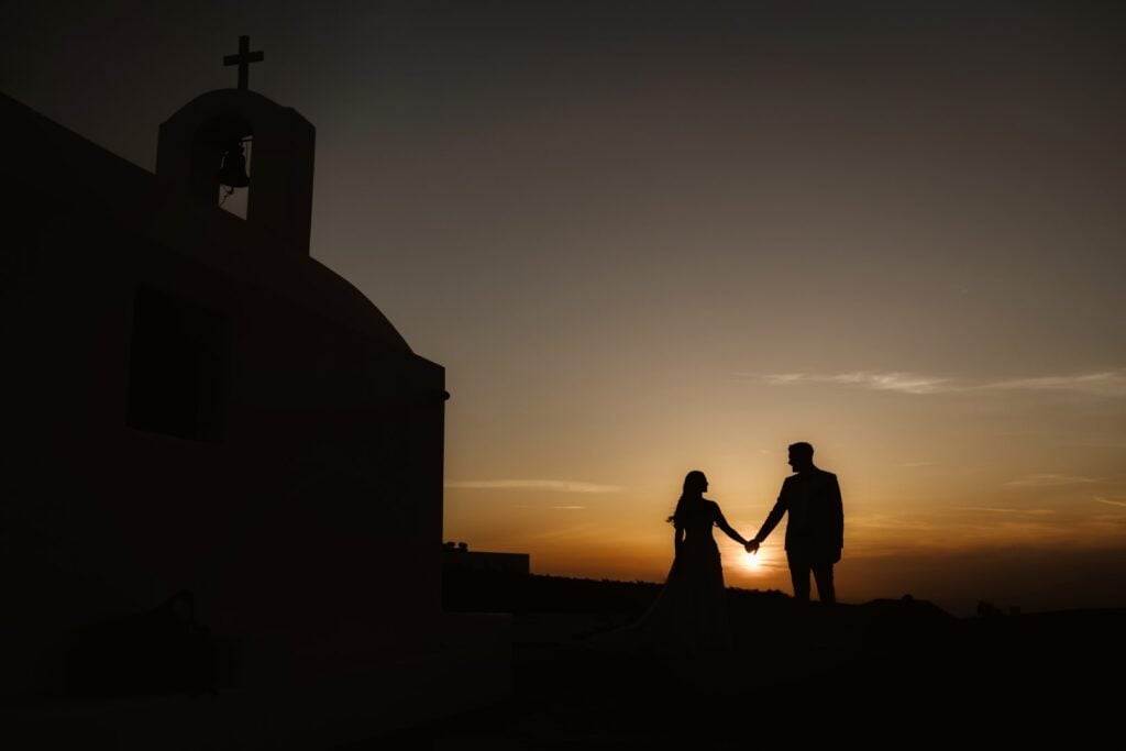 Silhouette of a couple holding hands at sunset near a church, framed by a warm, glowing sky—perfect for Destination Wedding Photography that captures unforgettable moments.