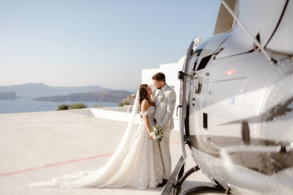 A bride and groom embrace beside a helicopter on a rooftop helipad, with a scenic coastline in the background—an unforgettable moment captured by a destination wedding photographer under the clear blue sky.