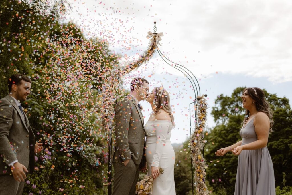 A couple share a kiss at an outdoor wedding ceremony under a decorated arch as colourful confetti rains down—captured beautifully by a Lake District Elopement Photographer, with guests smiling and trees framing the partly cloudy sky.