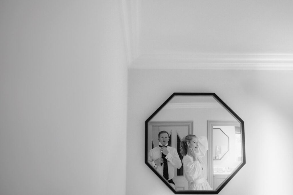 A black and white photo by a Lake District Elopement Photographer shows a man adjusting his tie and a woman fixing her hat, both reflected in an octagonal mirror on a white wall in a bright room.
