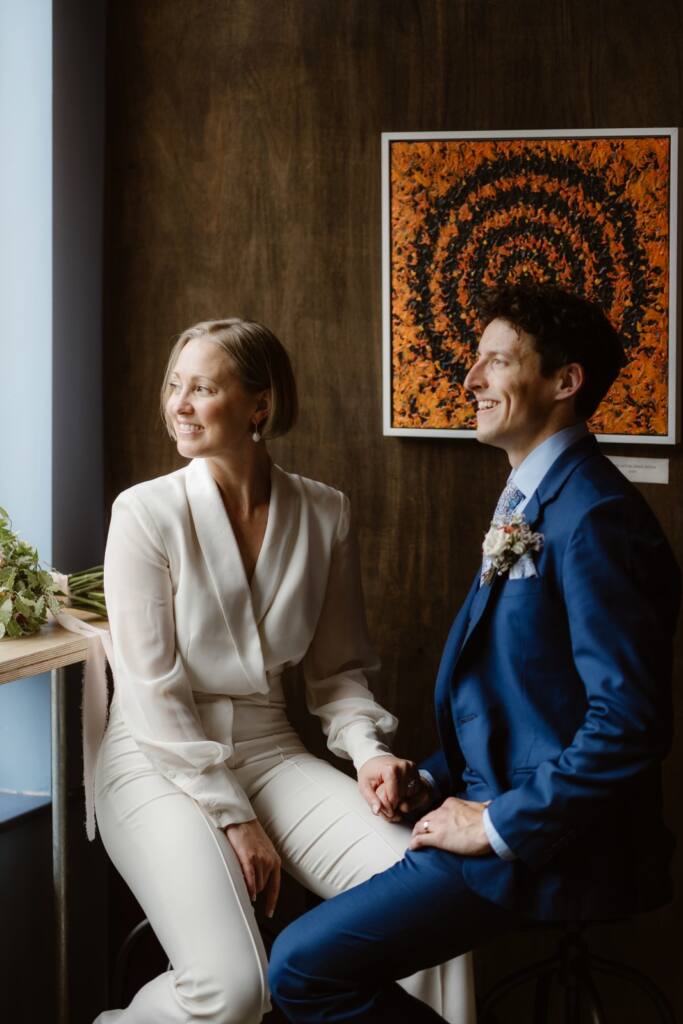 A couple dressed in formal attire smiles whilst sitting by a window. Captured by a Lake District Elopement Photographer, they hold hands and look towards the light, with a colourful abstract painting behind them.