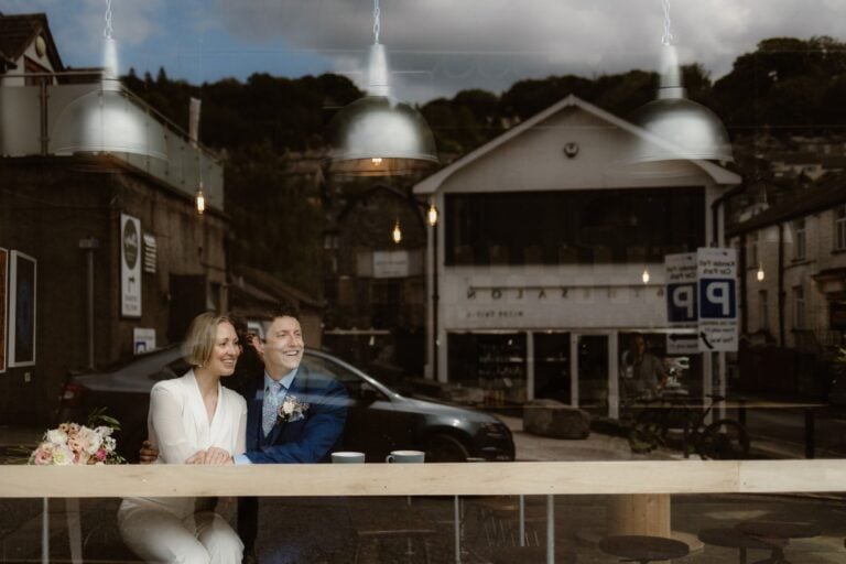 A smiling bride and groom sit together at a wooden counter inside a café, viewed through the window—captured by a Lake District Elopement Photographer. Street reflections, overhead lights, and a bouquet on the counter complete this intimate scene.