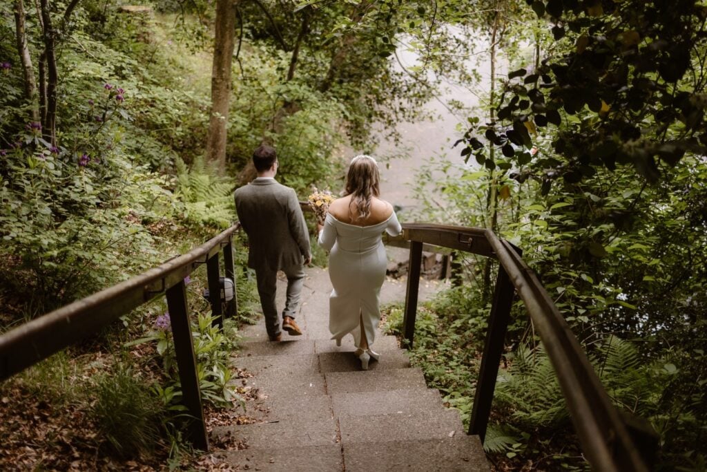 A couple in formal attire, the woman in a white dress, walk down a wooden stairway surrounded by lush greenery and trees—a perfect moment for a Lake District Elopement Photographer to capture in this enchanting wooded setting.