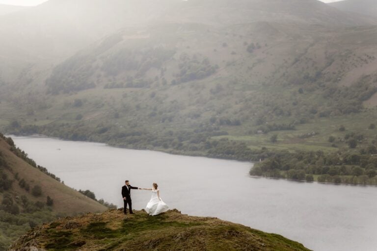 A bride and groom stand on a grassy hilltop, holding hands with a river and misty mountains in the background under soft, diffused light—perfect for a Lake District Elopement Photographer capturing unforgettable moments.