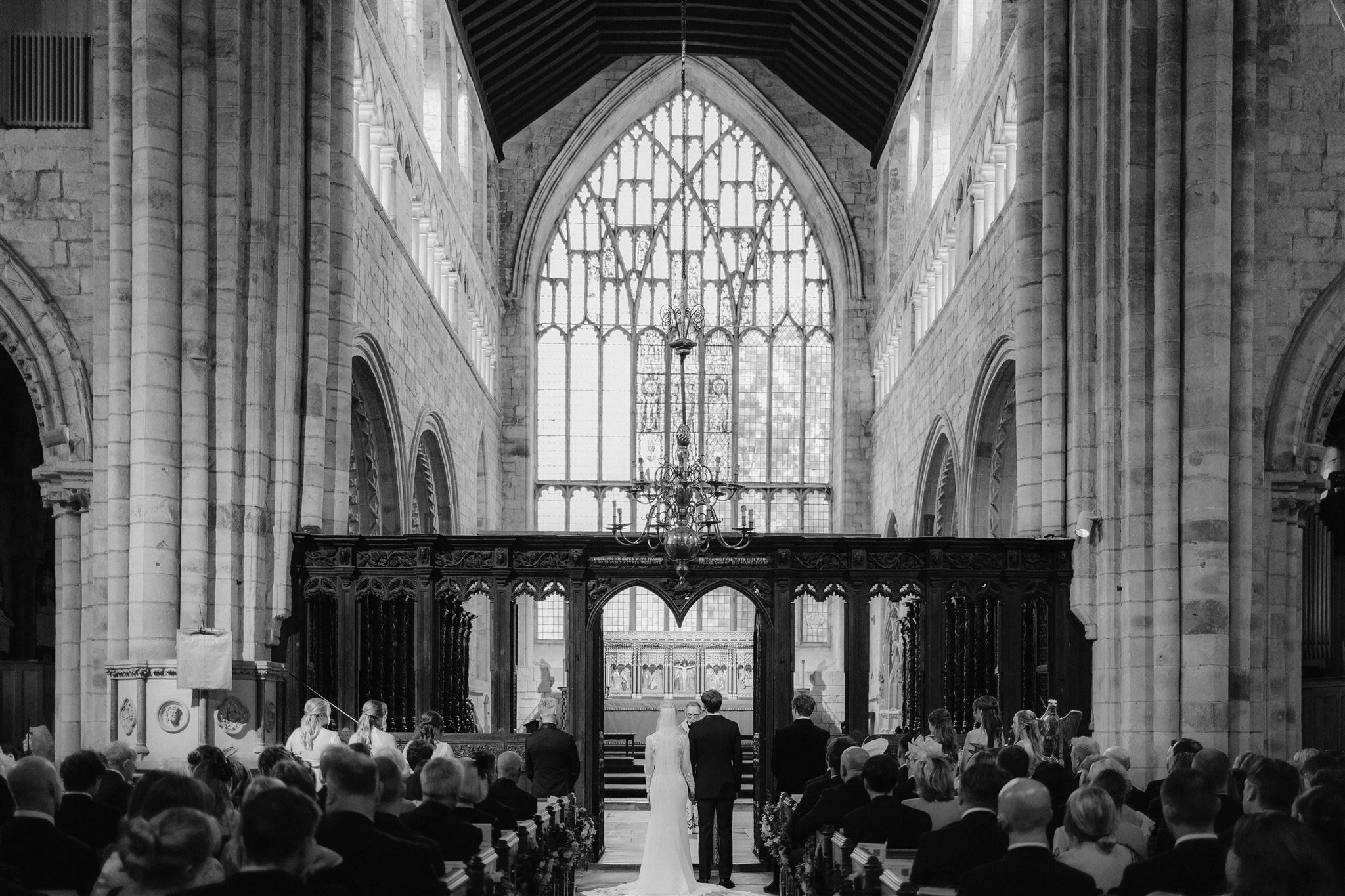 A bride and groom stand at the altar of a large, historic stone church, surrounded by seated guests. Tall arched windows and ornate architecture complete this timeless Holker Hall Wedding scene, captured in black and white.