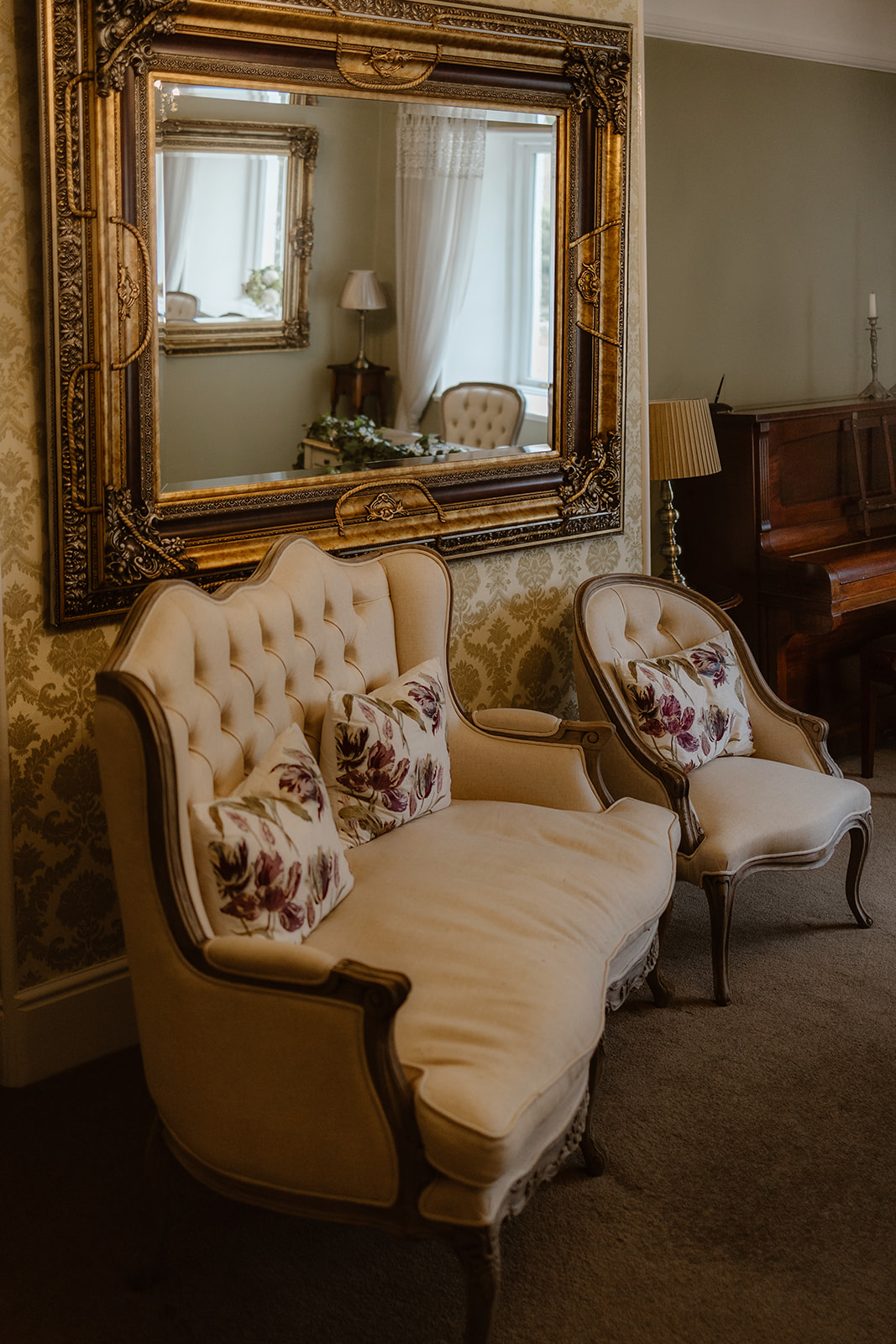 Vintage cream-coloured armchair and loveseat with floral cushions sit beneath an ornate gold-framed mirror in an elegant, softly lit room at Cote How, with patterned wallpaper and a piano in the background.