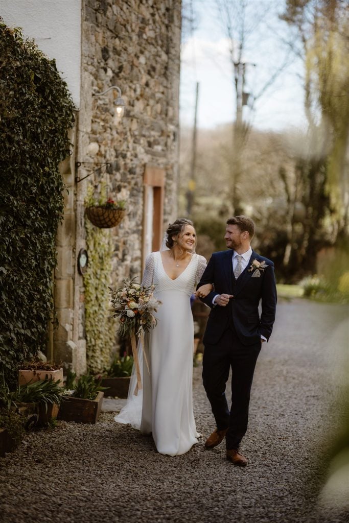 A bride in a white dress and a groom in a navy suit walk arm in arm, smiling at each other, along a gravel path beside Low Hall—a stone building covered in greenery. The bride holds a bunch of flowers.