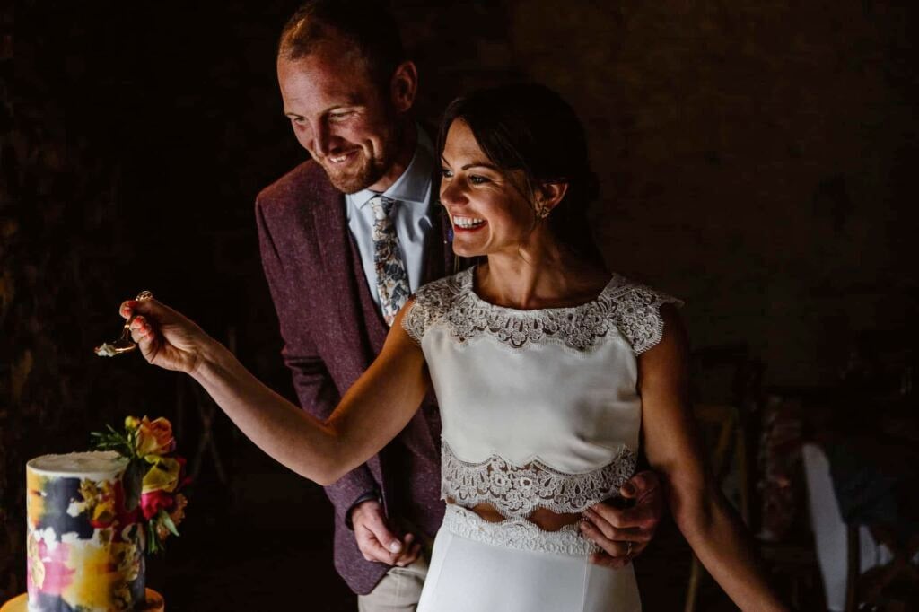 A smiling bride and groom stand together at Low Hall as the bride prepares to cut a colourful wedding cake, holding a piece of cake on a fork. The groom stands behind her, gently holding her waist.