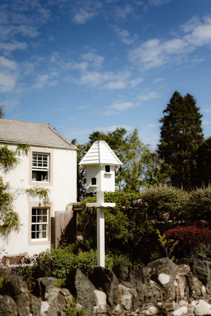 A white bird box on a tall post stands in a garden with greenery and flowers at Low Hall, in front of a white house with large windows, under a blue sky with scattered clouds.