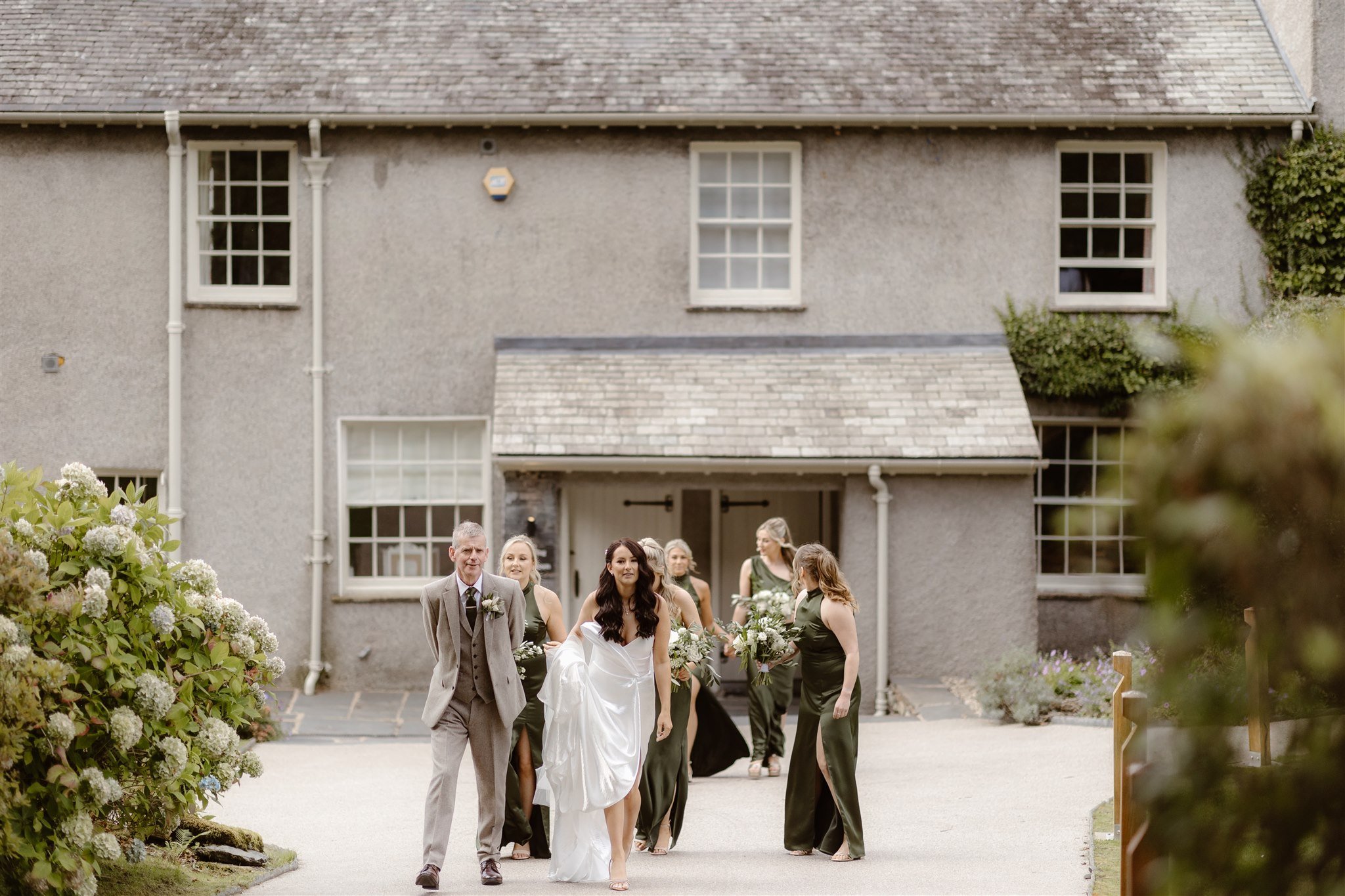 A bride in a white dress walks arm-in-arm with an older man in a suit, followed by bridesmaids in dark green dresses, outside the elegant Town Head Estate with its white-trimmed windows and flowering shrubs.