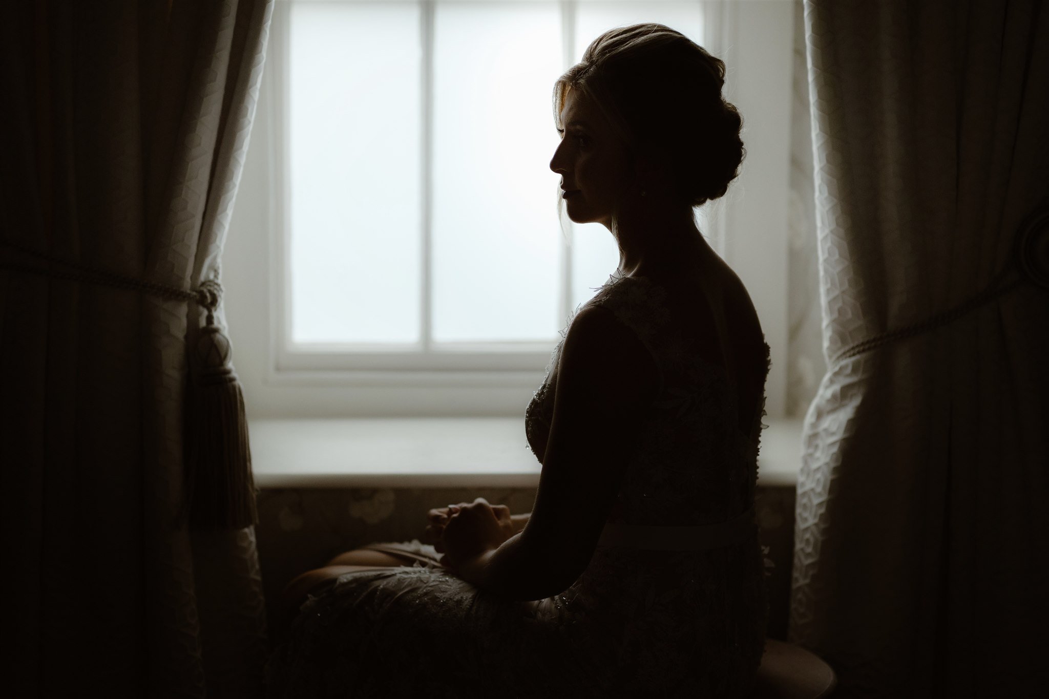 A woman in a dress sits in profile by a window with sheer curtains at Town Head Estate, softly lit from behind, creating a gentle, contemplative silhouette.